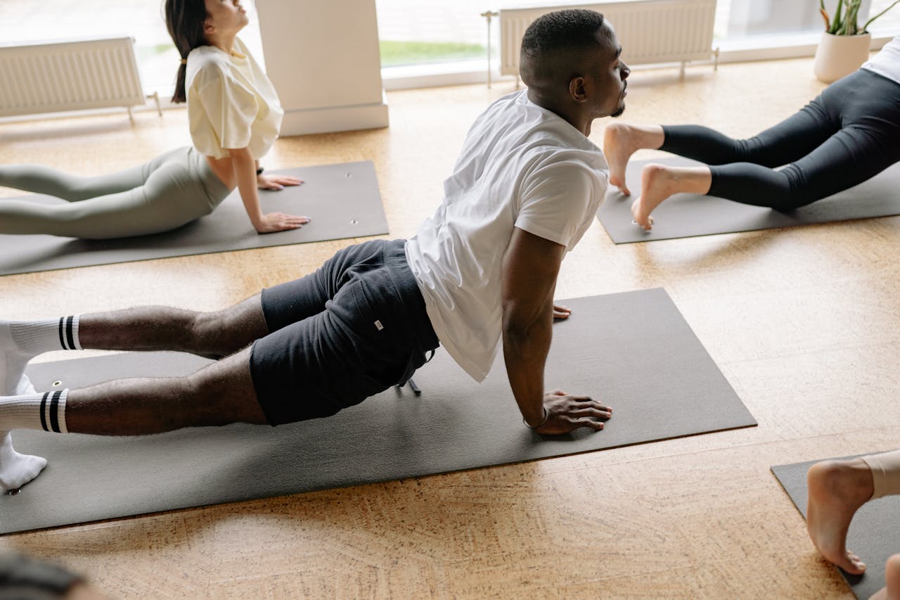 Adults in a yoga class performing stretches on mats in a well-lit studio.